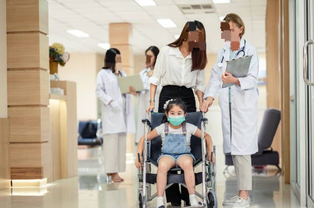 Smiling infant using a brightly colored wheelchair surrounded with the aid of present day pediatrician equipment.